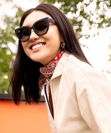 A woman smiles and wears the TOMS Traveler Sydney Matte Black sunglasses